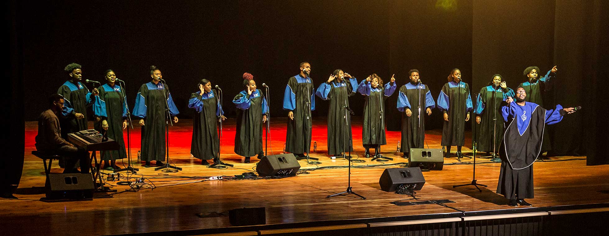 Twelve members of the Howard Gospel Choir perform on stage accompanied by a keyboardist and choir director. They all wear traditional gospel choir robes.