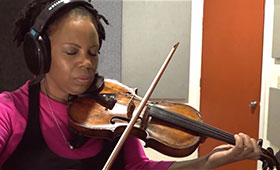 A woman in a recording booth closes her eyes and wears headphones as she plays a violin.