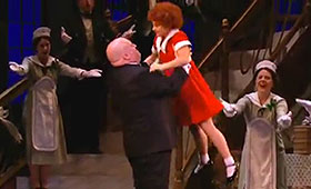 Servants hold their arms out in a welcoming gesture while Daddy Warbucks lifts Annie up into a hug.