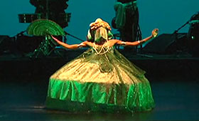 A female dancer, wearing a traditional Brazilian headdress and tent-style gown, outstretches her arms and legs while she holds a fan in her right hand during a folk dance.