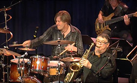 A man plays the drum while another man plays the saxophone to his right.