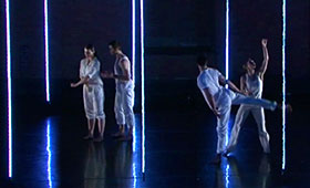 Dancers gesture while standing in front of floor-to-ceiling neon lighting.