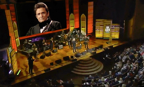 A group of musicians and singers on a stage in front of an audience perform below a large screen projecting an image of Johnny Cash.