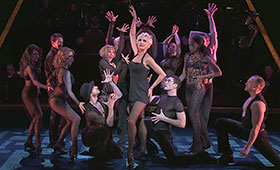 Roxie stands on stage with her arm in the air while surrounded by dancers.