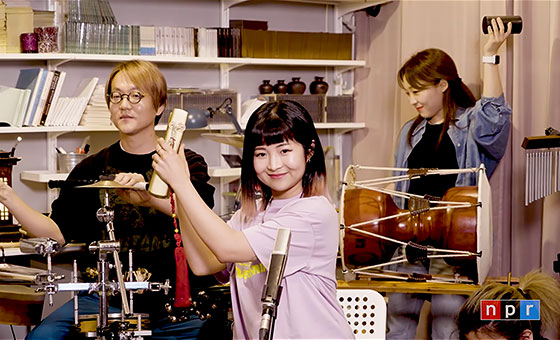 Three Korean musicians smile as they play traditional percussion instruments.