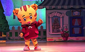 An actor dressed in an oversized Daniel Tiger costume makes a gesture with his left paw.
