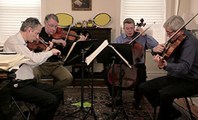 Four middle-aged gentlemen play violin and cello in a living room.