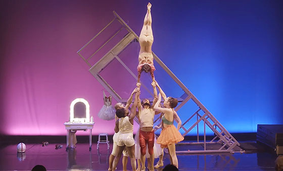 An acrobat in a handstand balances atop a group of acrobats standing in a base formation.