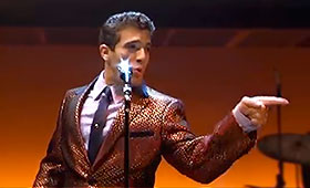 A man, wearing a shiny suit, sings in front of a microphone while pointing off stage.