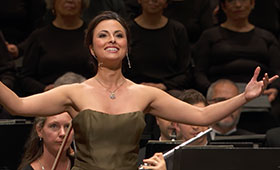 A woman stands in front of sitting musicians and outstretches her arms as she smiles at the audience.