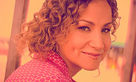 Joan Osborne sits in front of a window smiling with a patterned blouse and long curly hair.