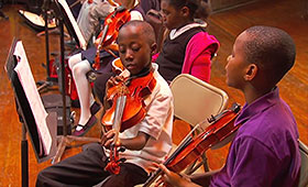 Two African-American boys sit in front of music stands and browse the parts of a violin.