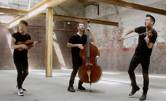 Three casually dressed musicians playfully perform their classical stringed instruments in a refurbished, open industrial space.