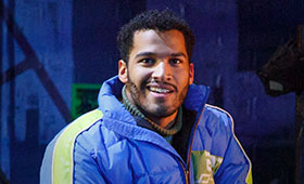 Christian Thompson wears a puffy winter coat on the stage of RENT.