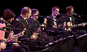 Five musicians sit in a row on stage while playing ukuleles.