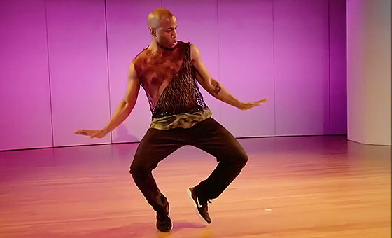 A man wearing loose clothing bends his legs at the knee while he stands on his toes and flares his hands out near his hips.