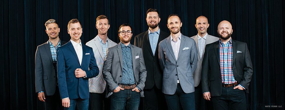 Eight male singers wearing sport jackets and button-up shirts smile for the camera.