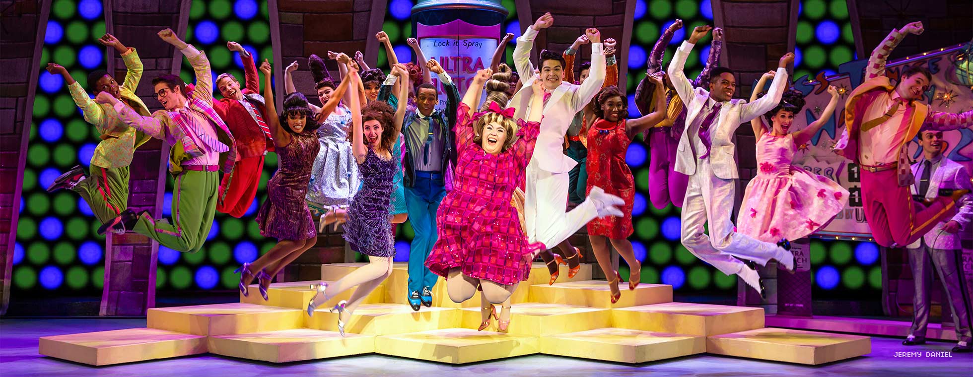 A girl wearing a beehive hairdo and a cast of dancers behind her jump up in unison.