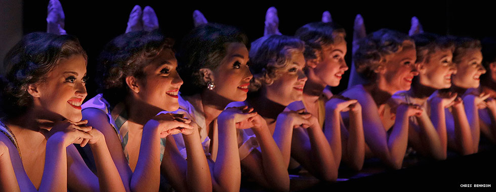Eight showgirls smile and prop their chins in their folded hands in a close-up photo.