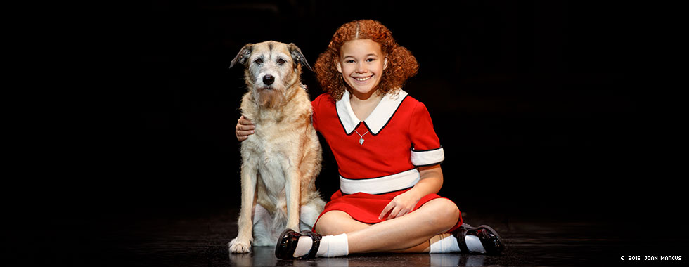 An actress portraying Annie smiles and sits cross-legged while she puts her right arm around Sandy the dog.