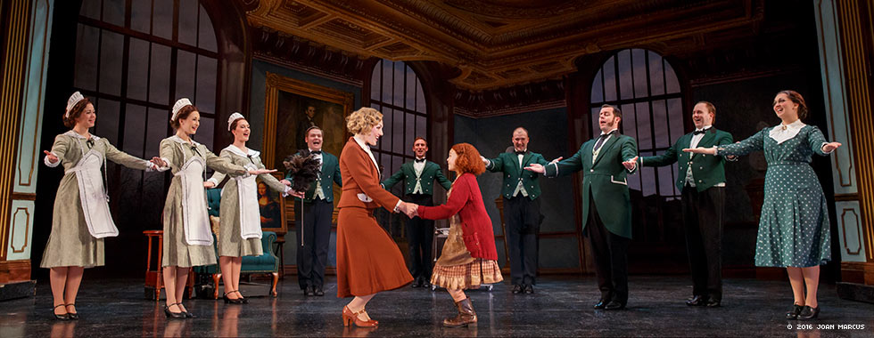 An actress portraying Annie smiles and holds both hands of an older actress while a staff of housekeepers and butlers with their arms outstretched surround them.