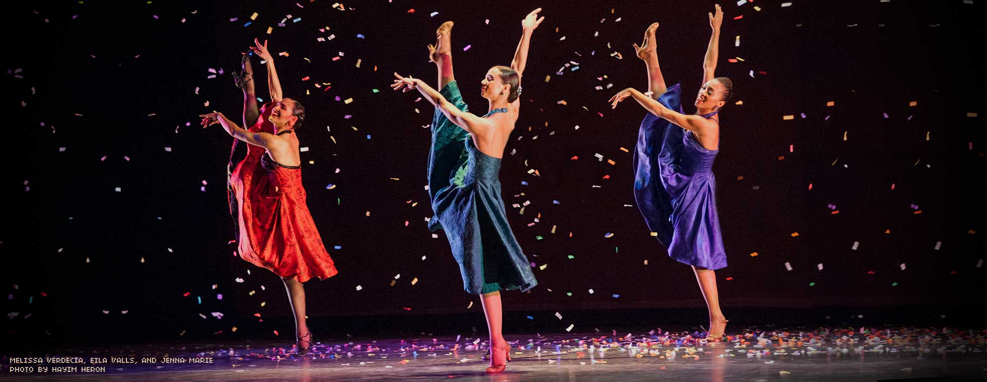 3 female dancers in traditional dresses each stretch with one leg in the air while standing on one foot. Confetti falls around them.