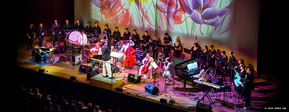 Chorus singers stand at the back of the stage behind Bang on a Can All-Stars and the ensemble's variety of stringed, percussion, and synthesizer instruments while a screen behind the musicians and singers show a painting of flowers.