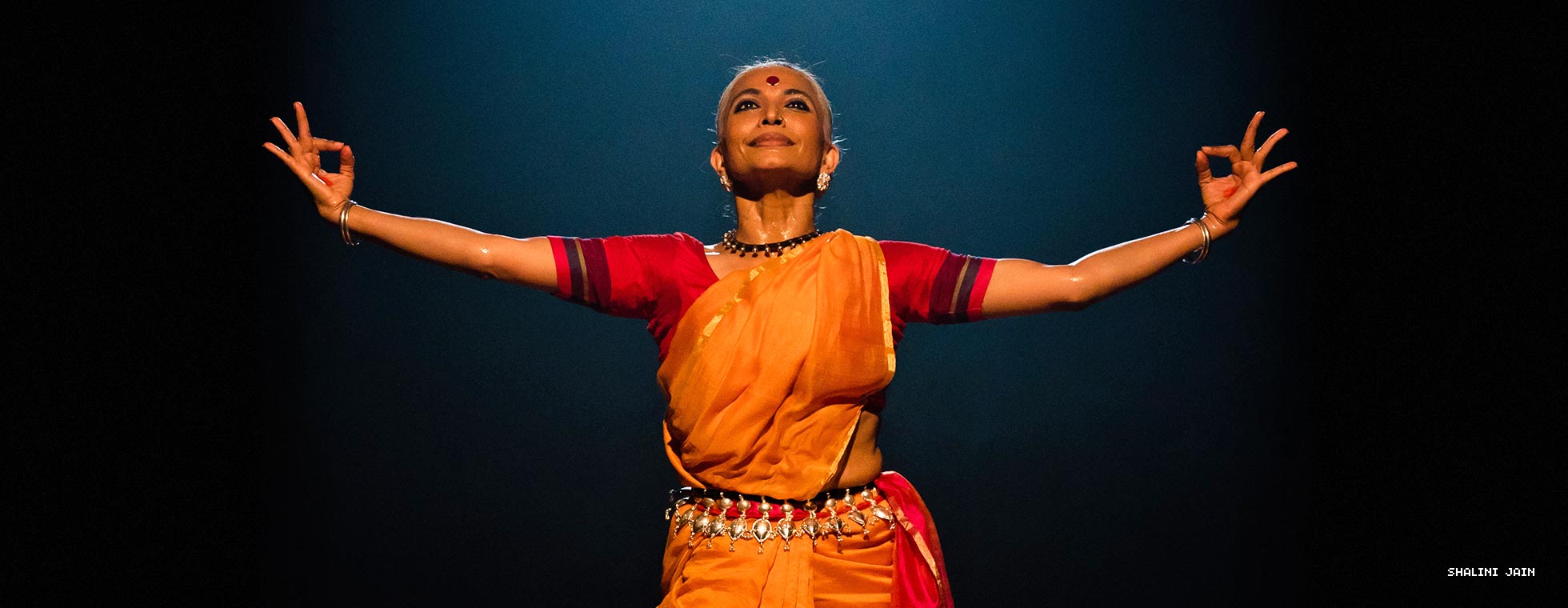 A classical Indian dancer smiles while she stands with her arms outstretched and her pointer fingers touching her thumbs.