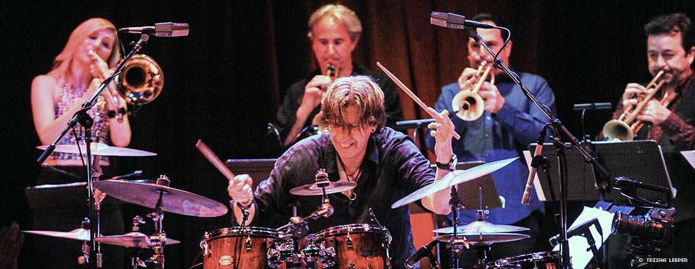 Drummer Tommy Igoe sits behind his kit and taps the cymbals while four brass musicians play behind him.