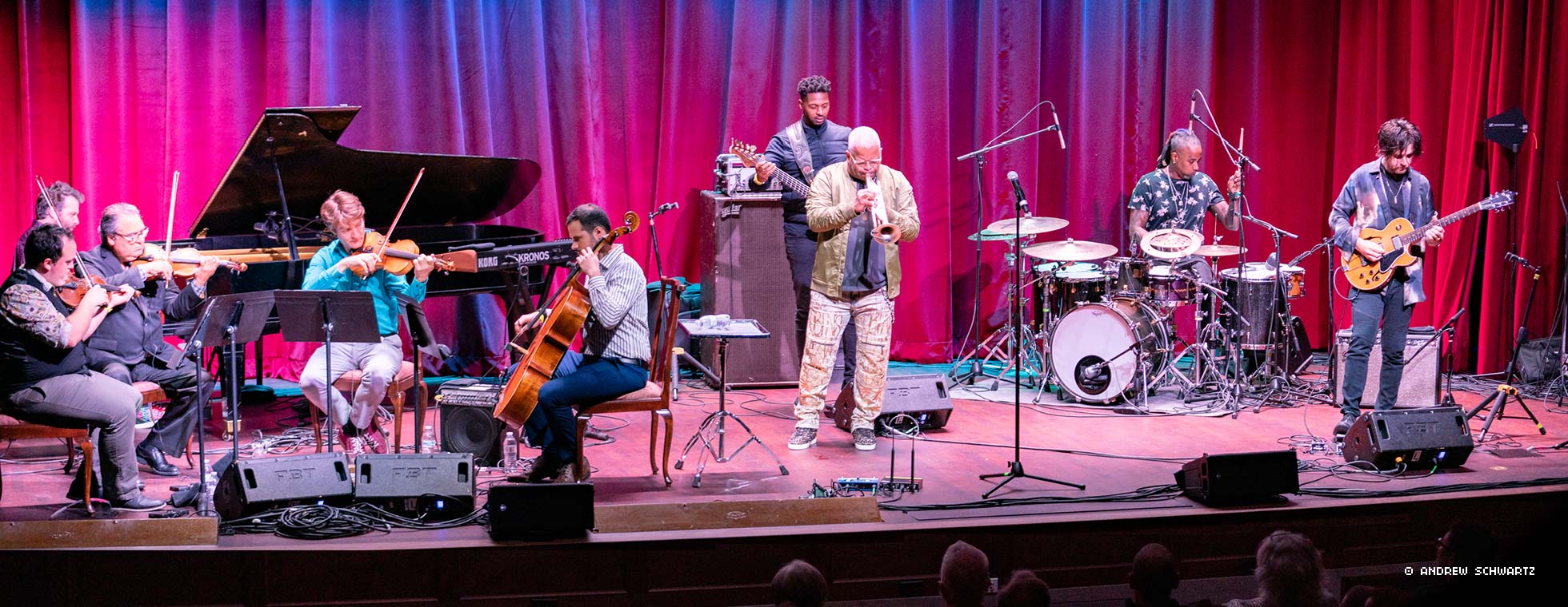 A diverse group of musicians—including a string quartet, electric guitar and synth players, a drummer, a pianist, and a trumpeter, gather together on a stage and perform.