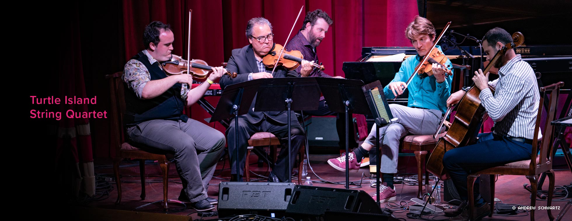 The Turtle Island String Quartet is shown sitting around a music stand and playing their instruments.
