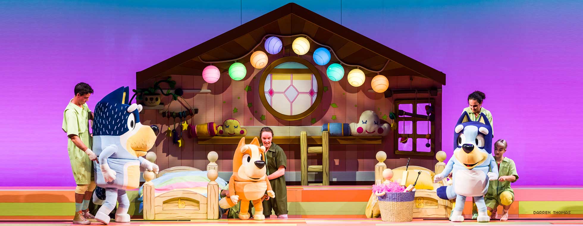 Actors stand behind three dog puppets to control their movements in scene depicting bedtime with a stage set with twin beds and children’s toys. 