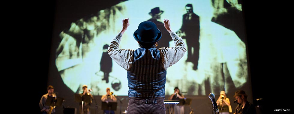 Darcy James Argue stands with his back to the crowd as he leads his Secret Society jazz ensemble in a performance while an illustration of two men shaking hands and a boy looking downward appears on a screen behind the ensemble.