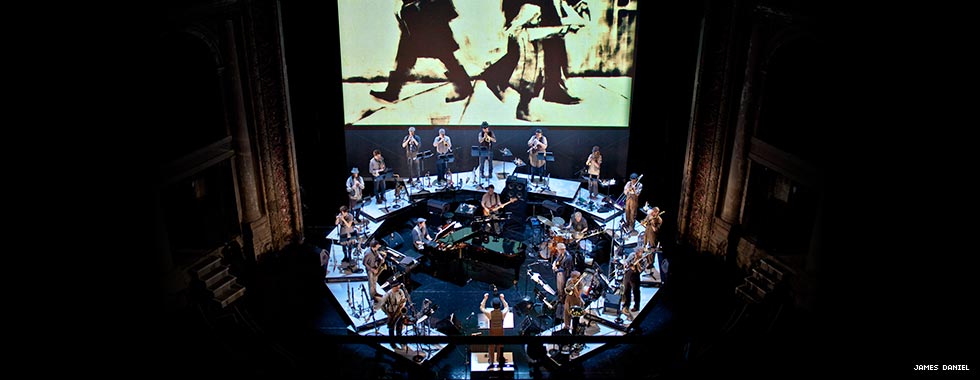 Darcy James Argue's stands with his back to the crowd as he leads his 20th-century-clothing-clad Secret Society jazz ensemble in a performance while a video depicting a child pointing to the right down appears on a screen behind the musicians who stand in a circle as they play.