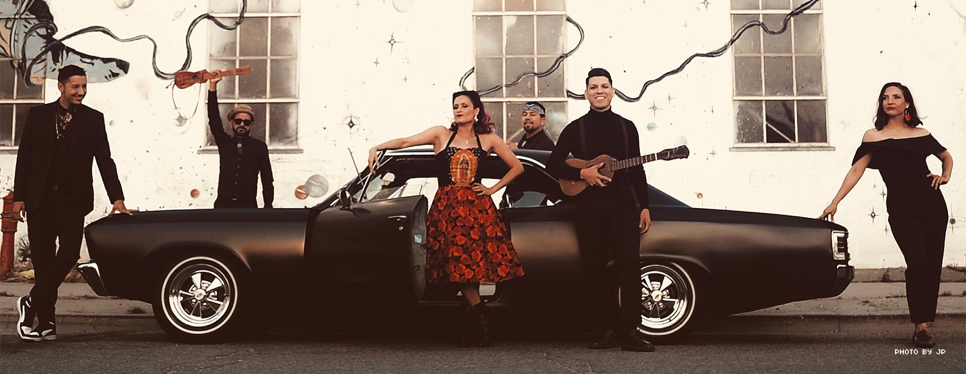 A band of glamorously dressed musicians—some holding stringed instruments—pose around and lean on a sleek, low-riding hot rod.