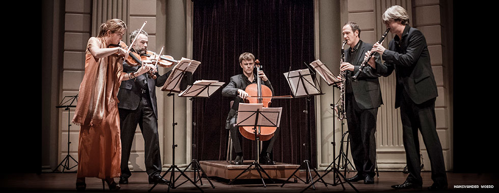 Four musicians, playing string and wind instruments, stand to each side of a man seated and playing a cello.