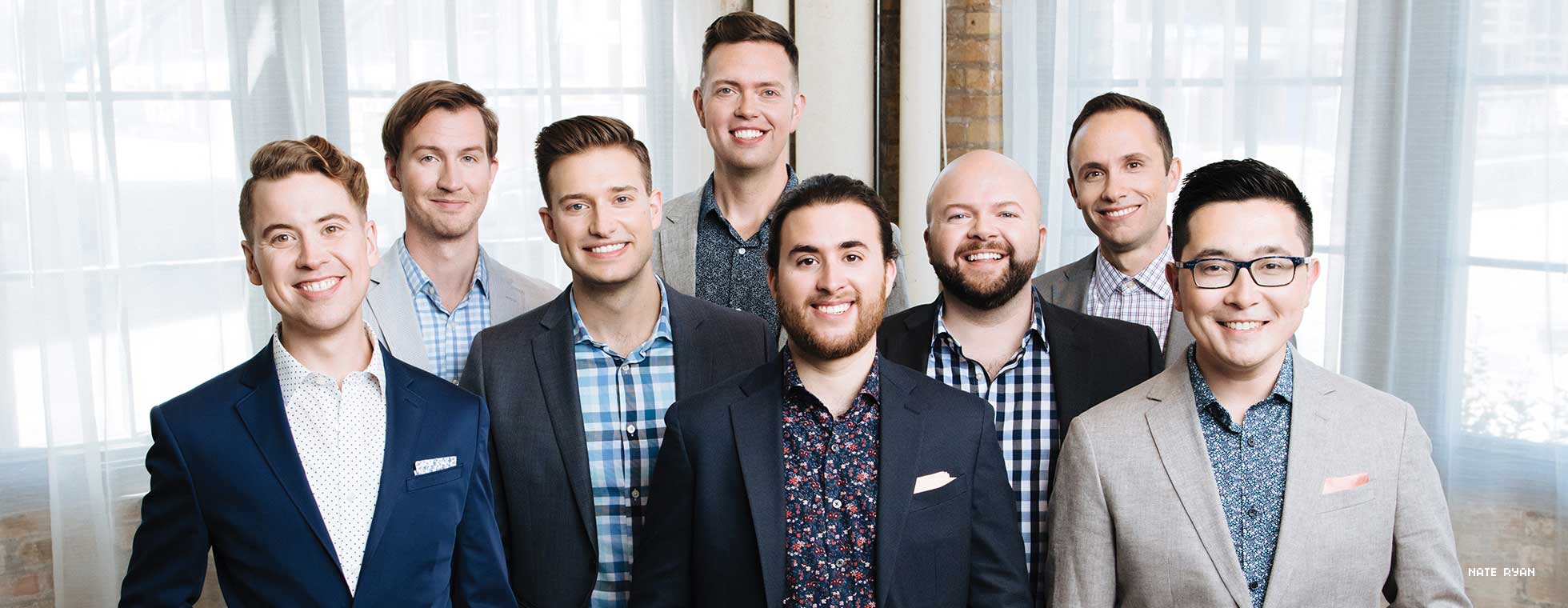 The eight members of Cantus stand together wearing casual suit jackets while smiling for the camera.