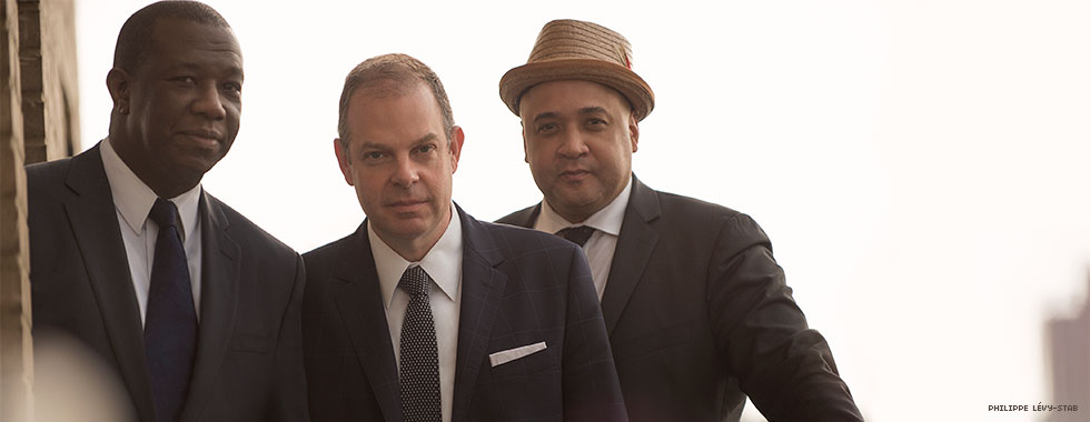 Three man in suits, with one wearing a homburg-style straw hat, look directly at the camera.