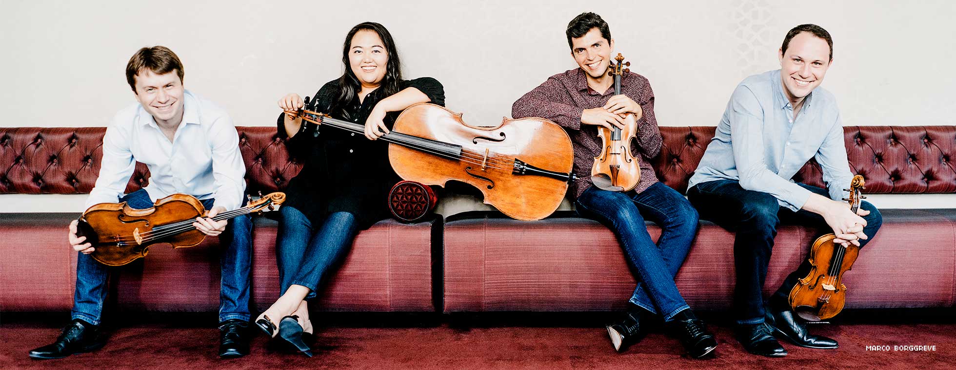 Four musicians sit casually on a couch with their instruments and smile.