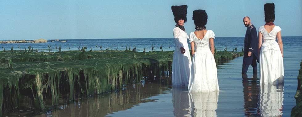 The members of DakhaBrakha, two of the musicians with their back to the camera and the remaining two looking over their shoulders to the photographer, wear traditional Ukrainian wedding attire and stand in seaside inlet with water reaching to their calves.