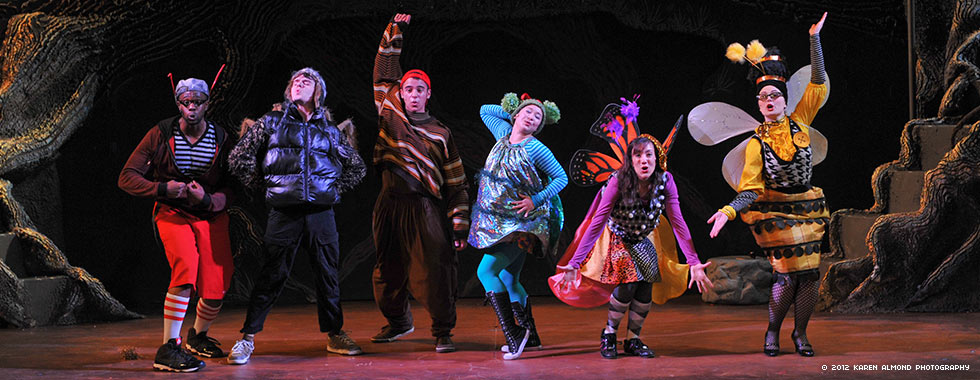 Six actors strike poses and make hand gestures while dressed in stylized insect costumes.