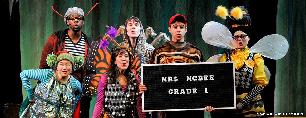 Actors dressed in costumes depicting a fly, beetle, butterfly, spider, worm, and bee hold a sign announcing Mrs. McBee’s first-grade class.