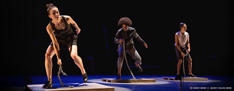 Three dancers stand bent over on boards as they extend a bent left leg back and drag a section of chain in on the floor with their right hands.