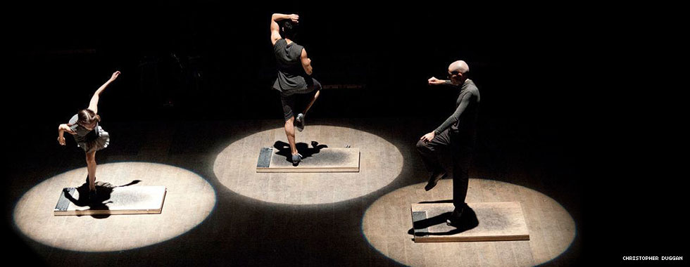 Spotlights illuminate three tap dancers each balancing on one leg on a board.