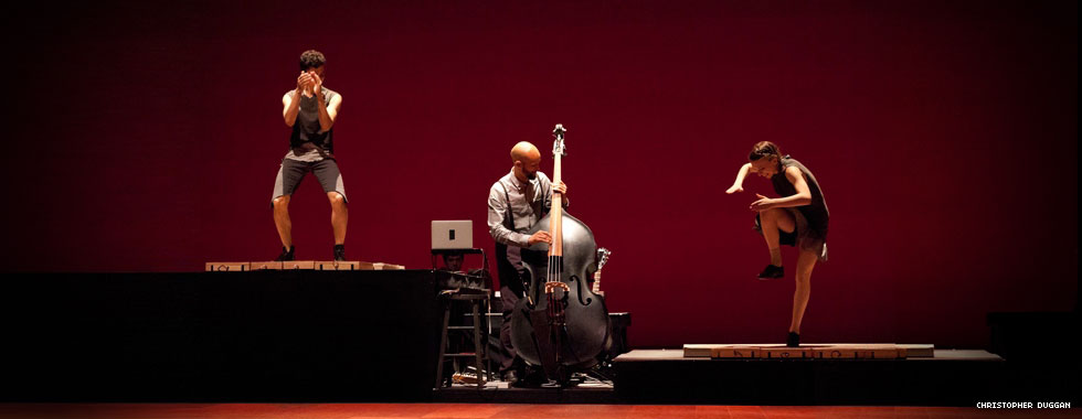 Two dancers on risers of different heights tap dance on each side of a cellist who plays his instrument.