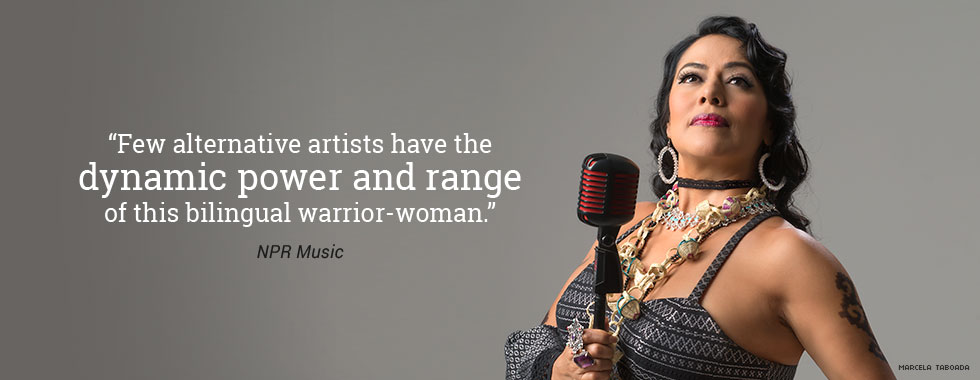 Lila Downs, wearing large baubles around her neck and on her fingers, holds a microphone in her right hand and looks up to the left.