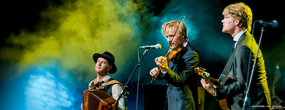 Three musicians—playing accordion, violin and cittern—perform on a stage while surrounded by stage fog.