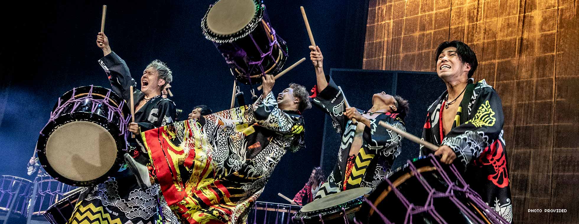 Men wear robes adorned with Japanese imagery as they bang sticks on large drums. 