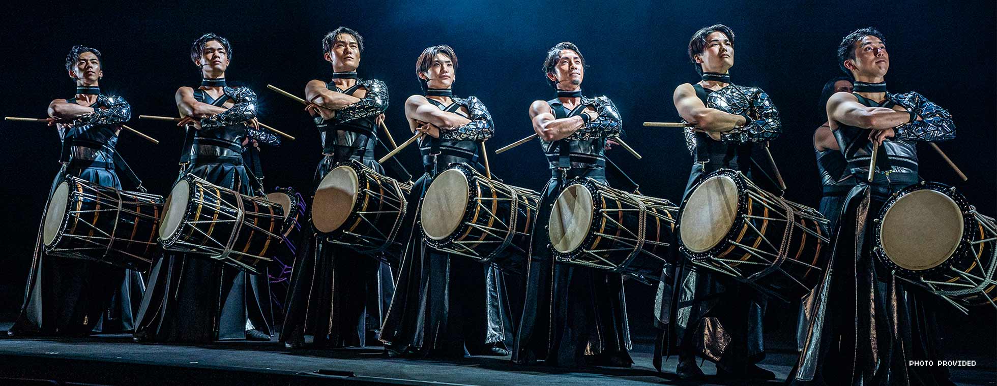 A group of Japanese men stand in a rigid line formation while drums hang from their torsos. 