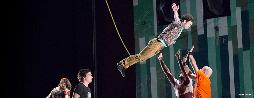 Two performers hold their hands up ready to catch a third acrobat who falls into their arms from a suspended rope.
